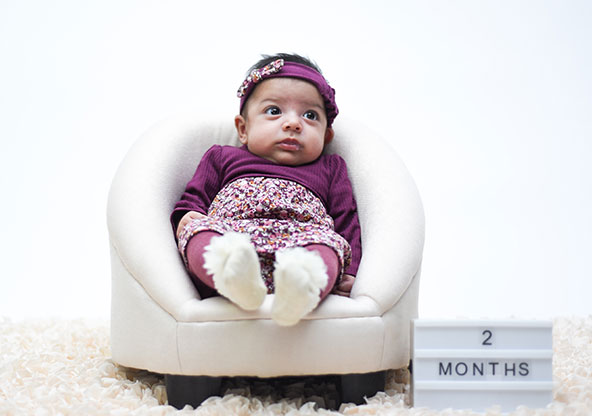 Baby Ana Isabella at 2 months old. She is sitting in a small chair, wearing a pink outfit and a pink headband.
